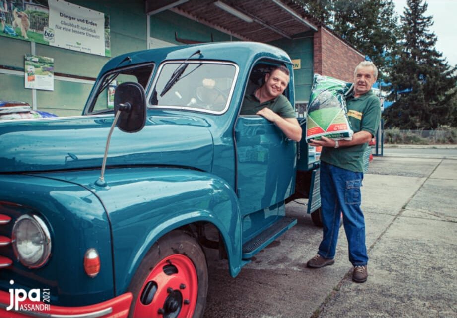 Ein Mann sitzt lachend in einem blauen Oldtimer-Lkw, während ein anderer daneben steht und einen Sack Frömmig Rasendünger Spezial hält. Beide tragen grüne Firmenkleidung. Im Hintergrund ist das Geschäftsgelände von Frömmig’s Gartenbedarf zu sehen.