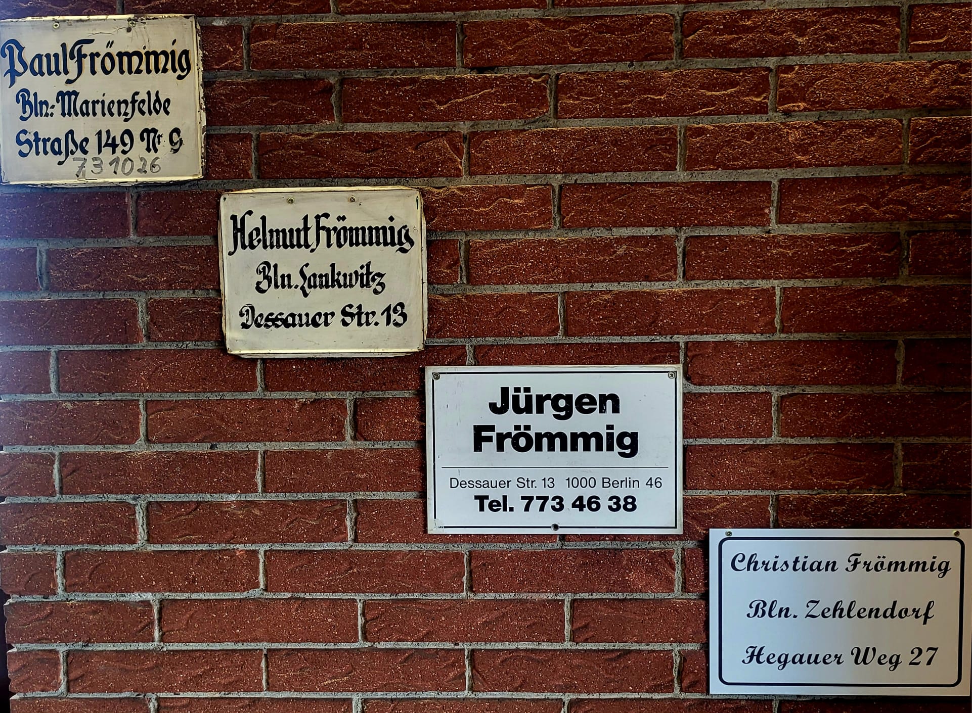 Fünf Namensschilder der Familie Frömmig an einer roten Backsteinwand. Die Schilder tragen die Namen Paul, Helmut, Jürgen und Christian Frömmig mit jeweiligen Berliner Adressen.